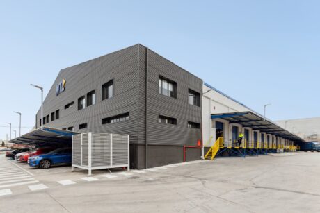 Nave industrial moderna con fachada de metal corrugado, múltiples muelles de carga, coches aparcados y dos trabajadores en una escalera.