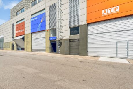 Fila de naves industriales cerradas con letreros de colores y puertas metálicas enrollables a lo largo de una calle asfaltada en un día despejado.
