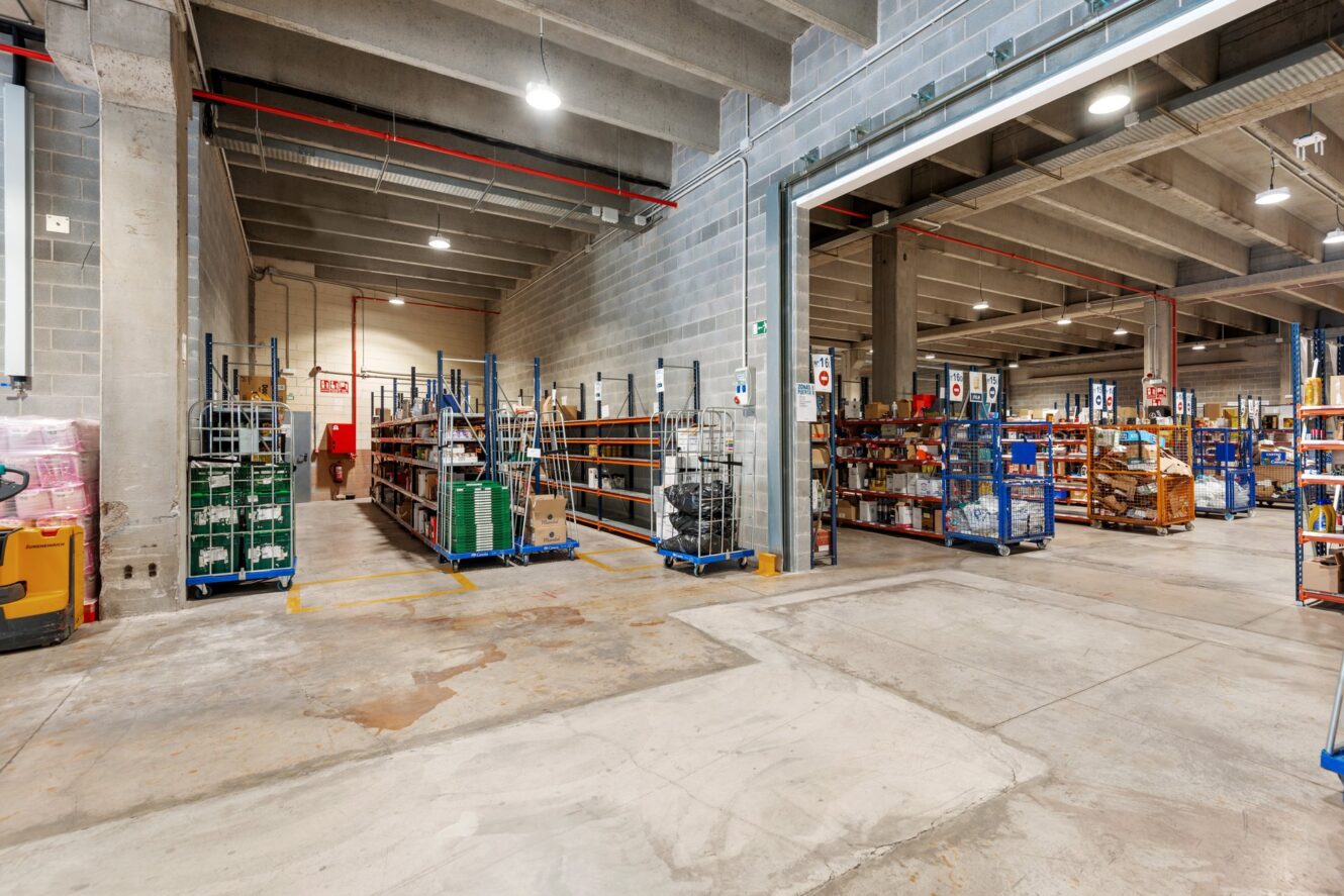 Interior de almacén espacioso con suelos de hormigón, techos a la vista y estanterías repletas de diversos productos empaquetados y en cajas. Se ven algunas estanterías independientes y una transpaleta.