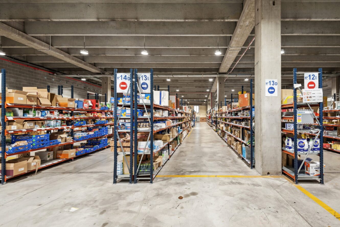 Pasillo ancho en un almacén con estanterías altas repletas de cajas y productos diversos, suelos de hormigón e iluminación superior visible.