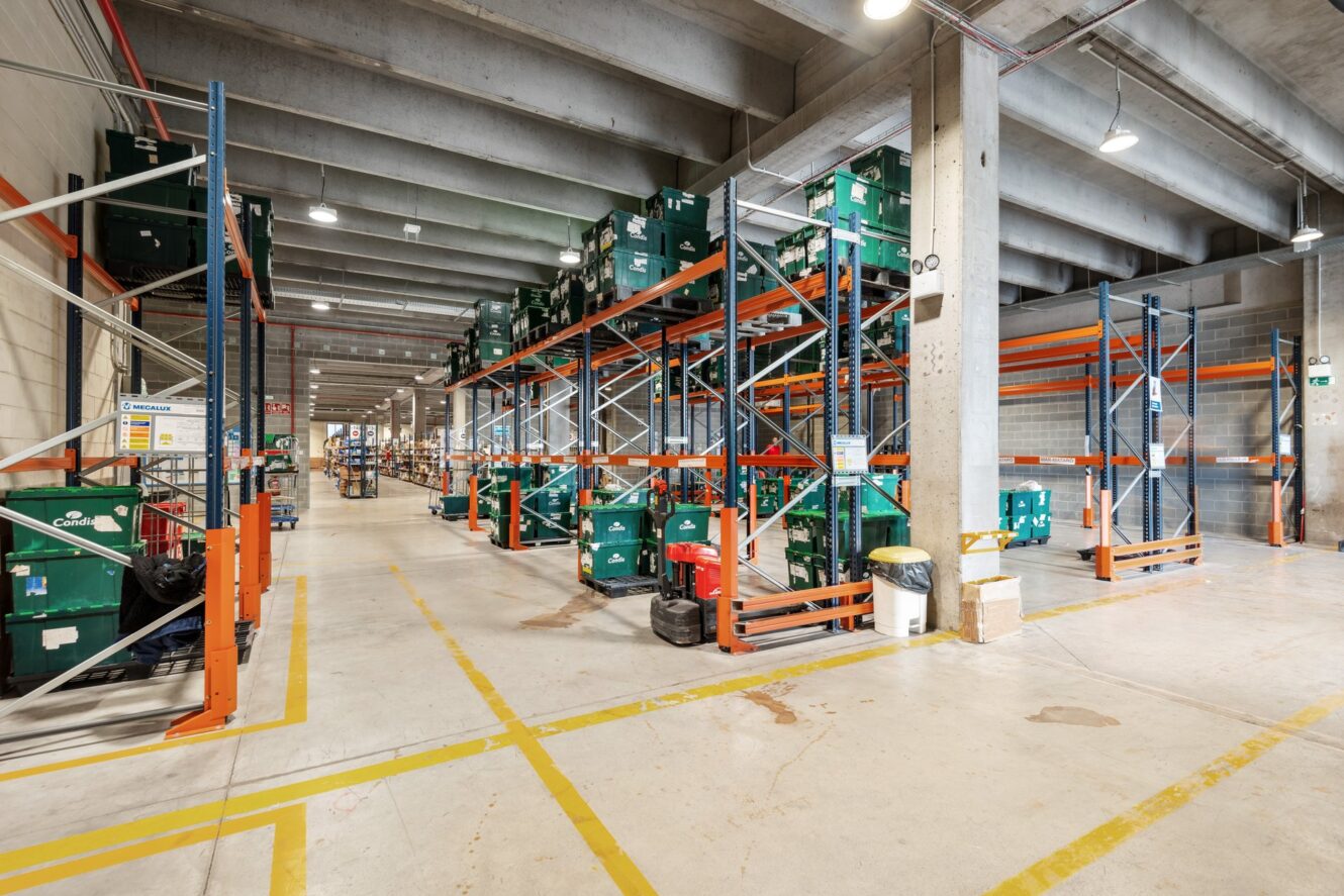 Interior de almacén con estanterías altas que sostienen contenedores de plástico verde, pilares de hormigón, techo alto y líneas amarillas marcadas en el suelo.