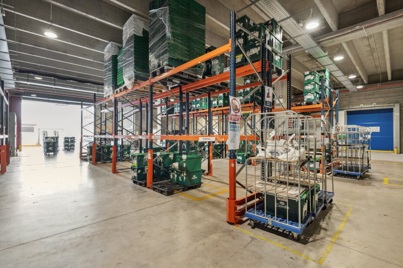 Interior de almacén con estanterías de palés, cajas apiladas, carros y equipos sobre un suelo de hormigón bajo un techo alto con tuberías a la vista.