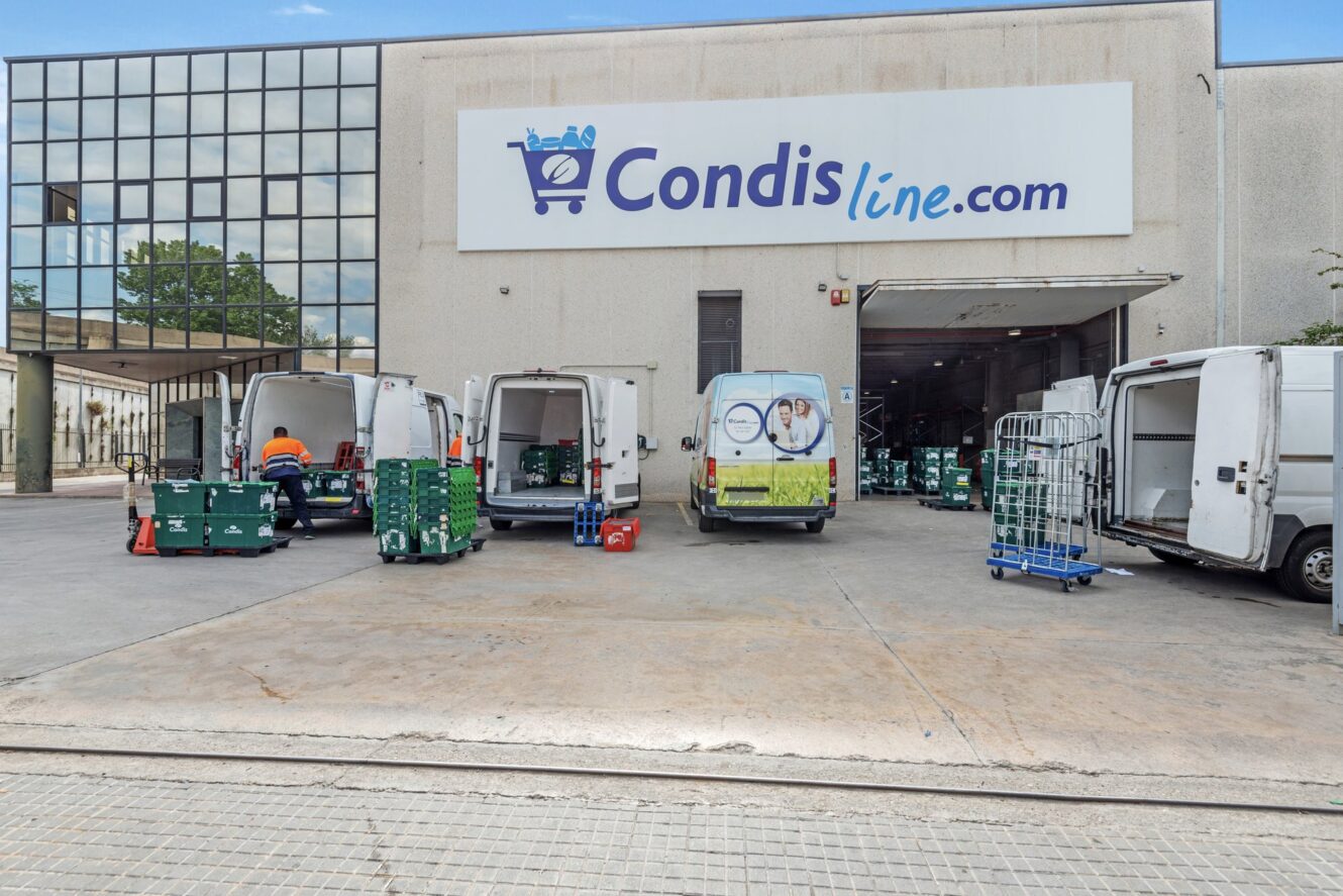 Varias furgonetas de reparto están aparcadas fuera de un almacén de Condisline.com, con trabajadores cargando y descargando cajas verdes y carros metálicos.