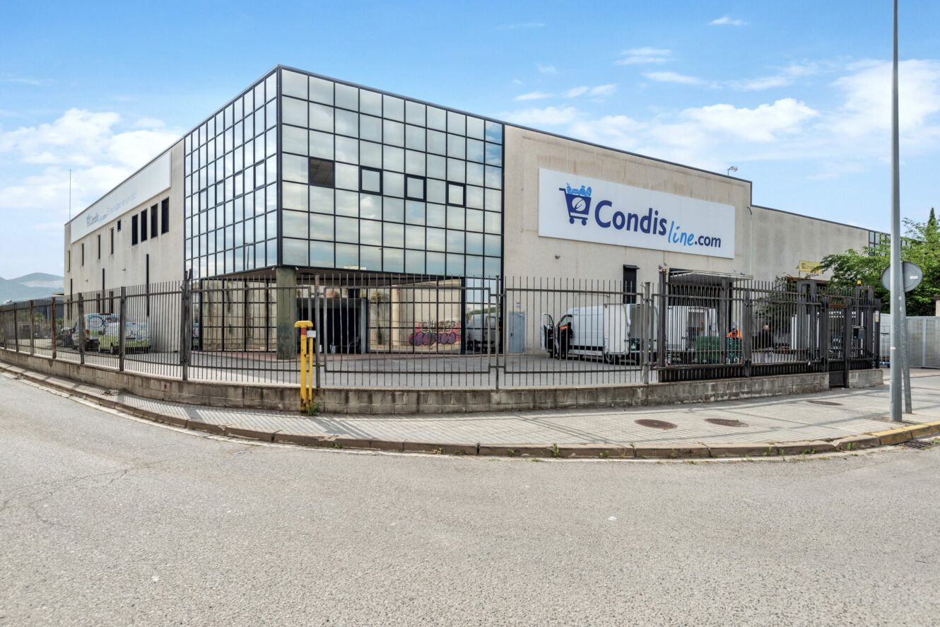 Un edificio comercial con un cartel de Condisline.com, fachada acristalada, perímetro vallado y furgonetas de reparto aparcadas dentro de la zona vallada.