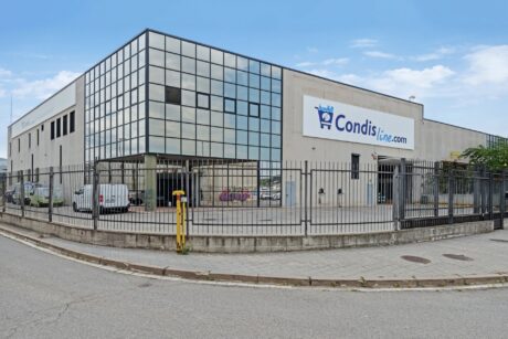 Un gran edificio comercial con ventanas de cristal y un cartel en el que se lee Condisline.com, rodeado por una valla metálica a lo largo de una calle en un día nublado.