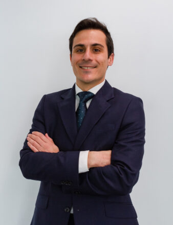 Hombre con traje oscuro y corbata, de pie sobre un fondo blanco liso, con los brazos cruzados, sonriendo a la cámara.