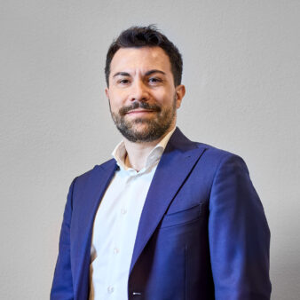 Un uomo con capelli corti e scuri e barba indossa una camicia bianca e una giacca da abito blu, in piedi su uno sfondo chiaro.