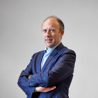 Un uomo in abito blu navy sta in piedi con le braccia incrociate, guardando la telecamera su uno sfondo chiaro.