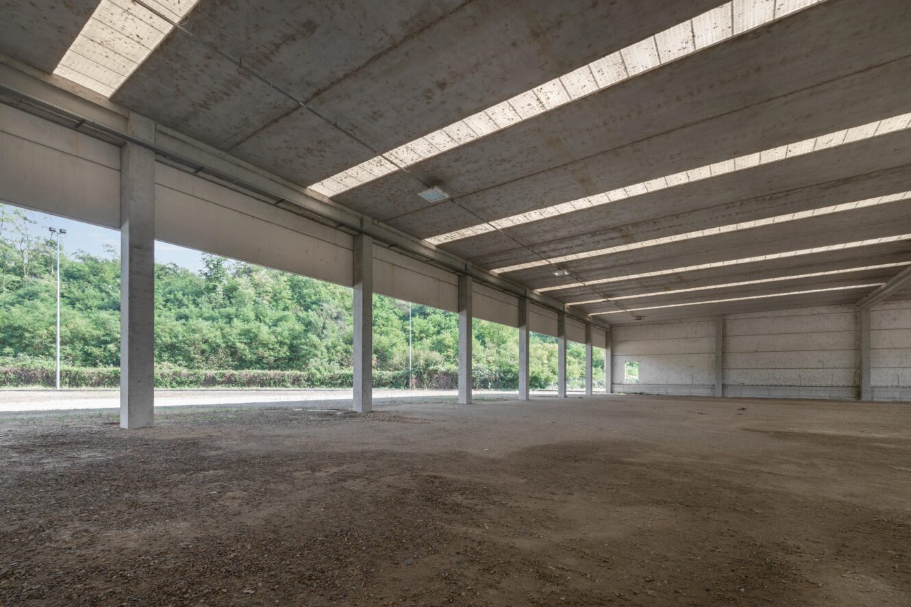 Interno di un grande magazzino industriale vuoto con pavimenti in cemento, colonne di sostegno e pareti parzialmente aperte; all'esterno sono visibili degli alberi.