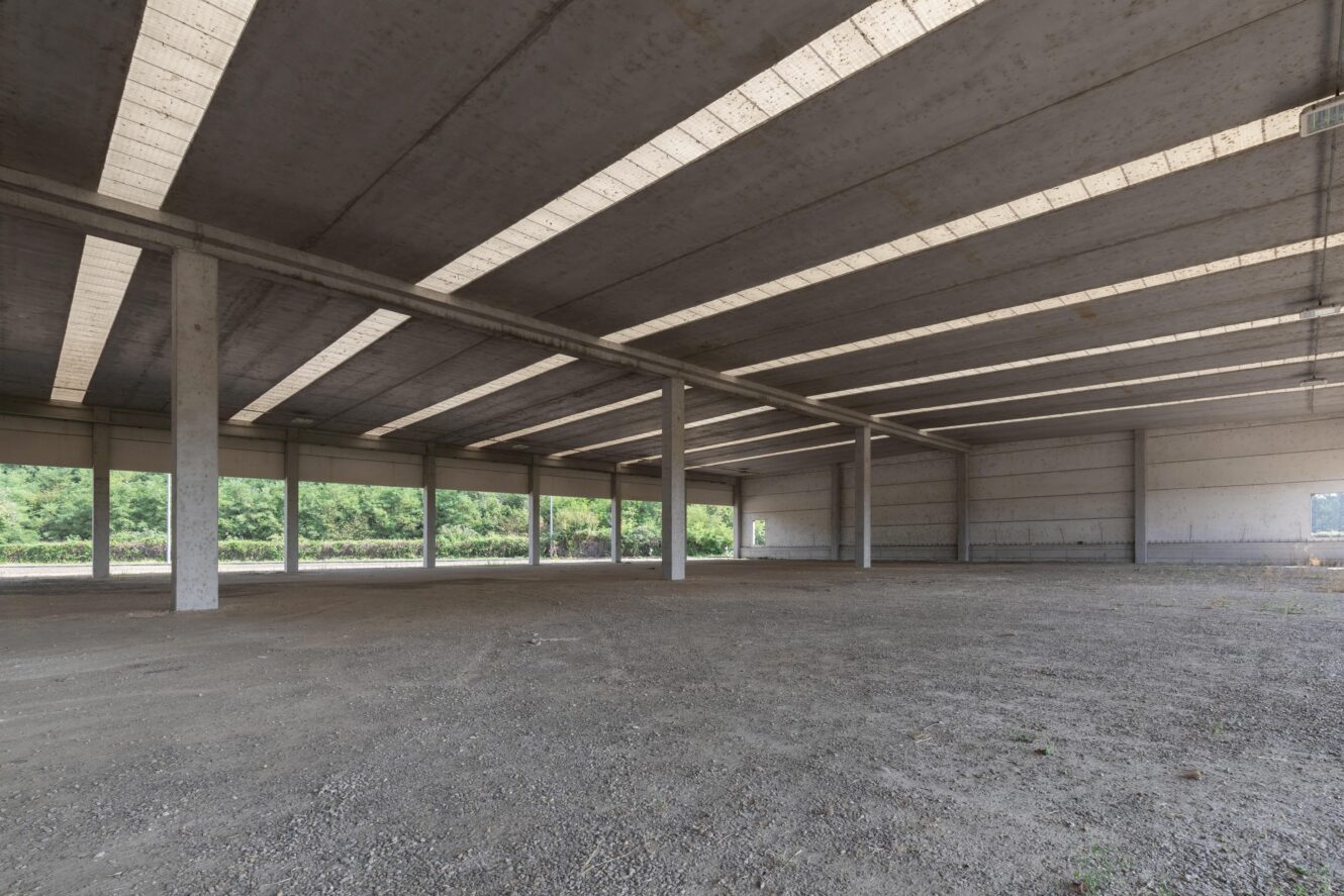 Magazzino industriale vuoto con pilastri in cemento, soffitto a vista e pavimento in ghiaia. Grandi finestre lungo le pareti lasciano entrare la luce naturale. Il verde è visibile all'esterno.