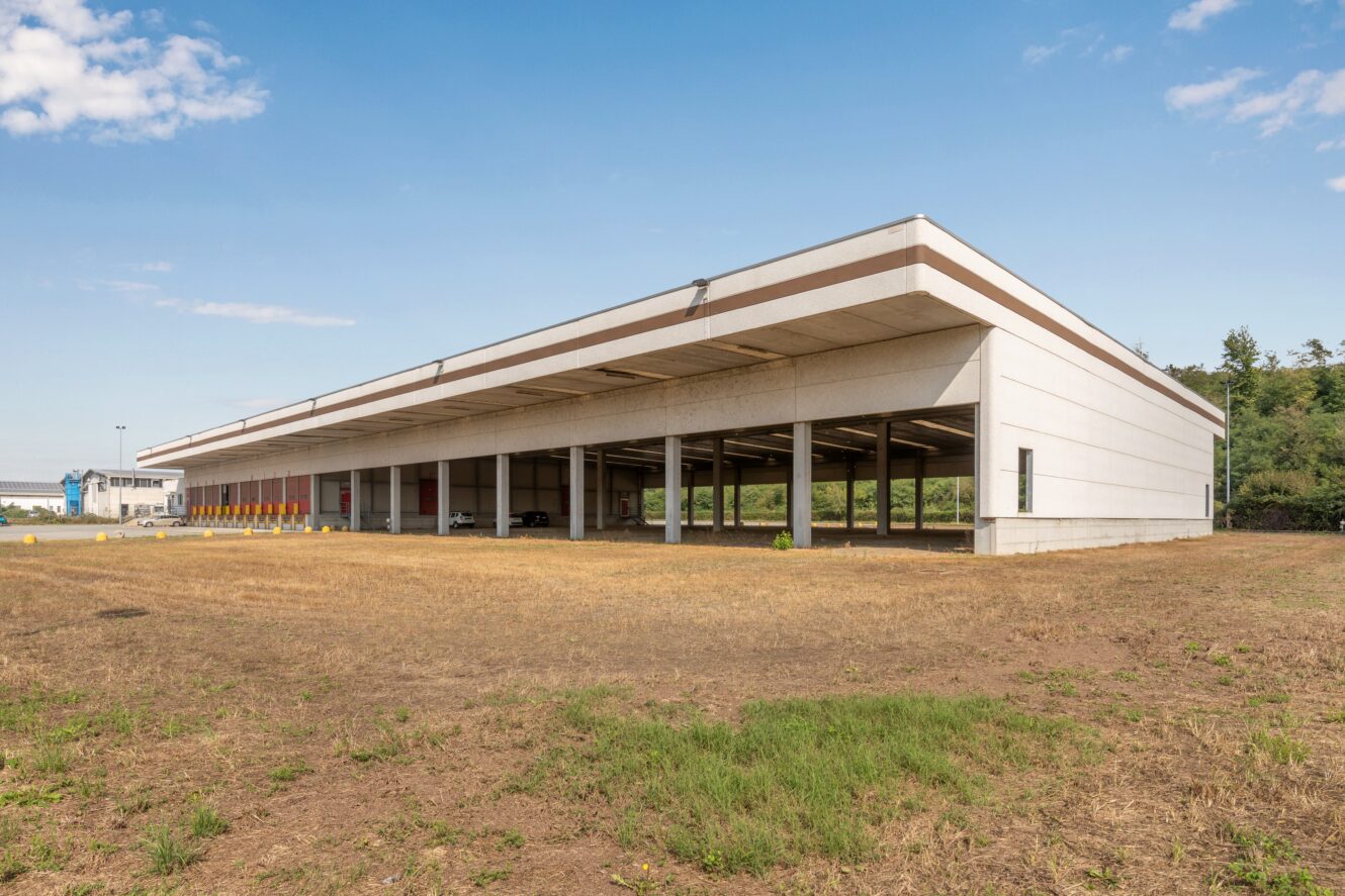 Un grande capannone industriale aperto, con banchine di carico e veicoli parcheggiati sotto, su un terreno erboso e asciutto, sotto un cielo limpido.
