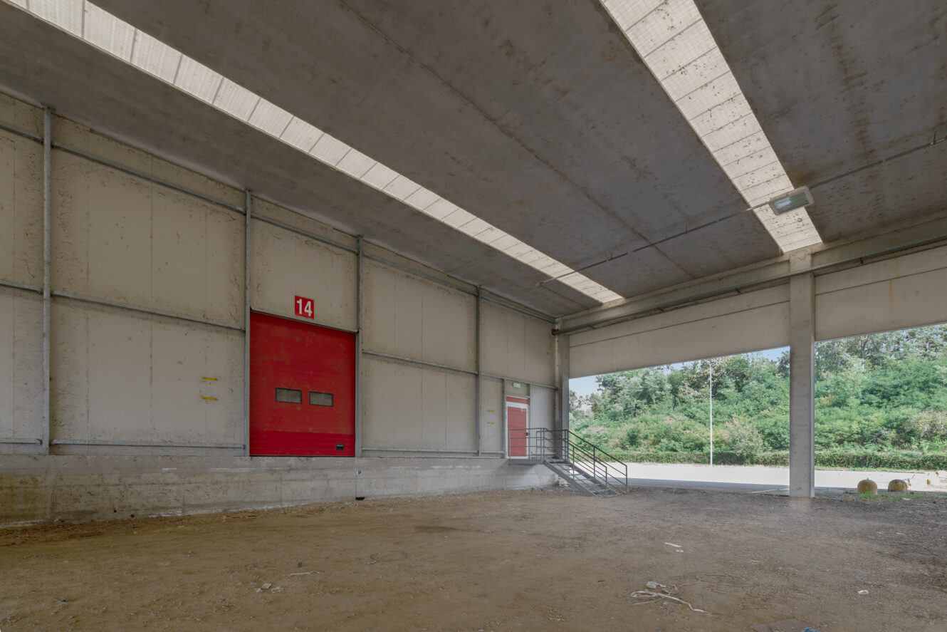 Un magazzino industriale vuoto con un'ampia entrata aperta, pavimenti in cemento, soffitti alti e una porta di carico rossa con l'etichetta 14. Alcuni detriti sono sparsi sul terreno.