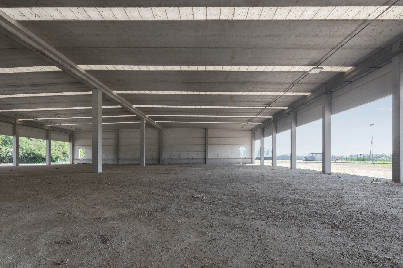 Interno di un magazzino di cemento vuoto e non finito, con pavimento in terra battuta, colonne di sostegno e grandi finestre che lasciano entrare la luce naturale.