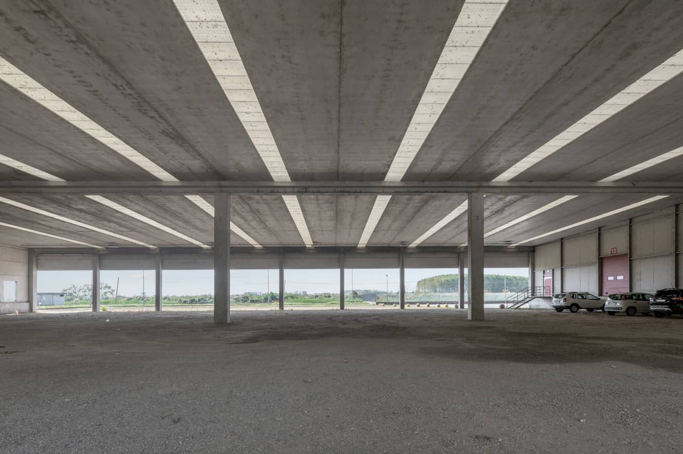 Un grande magazzino industriale vuoto con pavimenti in cemento, ampi pilastri, pareti aperte e alcune auto parcheggiate all'esterno. La luce naturale entra dal tetto e dalle aperture delle pareti.