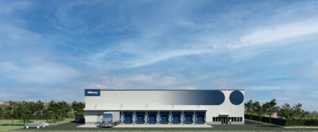 Un grande e moderno edificio adibito a magazzino con diverse banchine di carico, un camion per le consegne, auto parcheggiate e il verde circostante sotto un cielo azzurro.