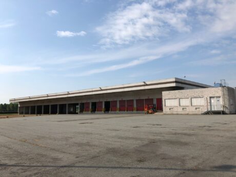 Una grande banchina di carico industriale vuota con diverse porte rosse, un ufficio bianco sulla destra e un ampio terreno asfaltato sotto un cielo parzialmente nuvoloso.