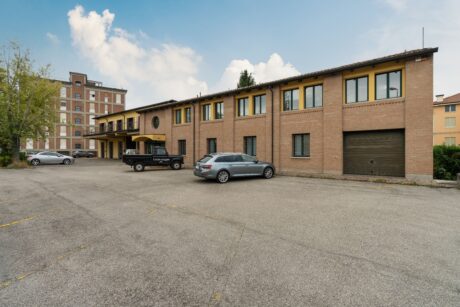 Un edificio in mattoni a due piani con più finestre e una porta di garage, diverse auto parcheggiate e un parcheggio asfaltato in primo piano.