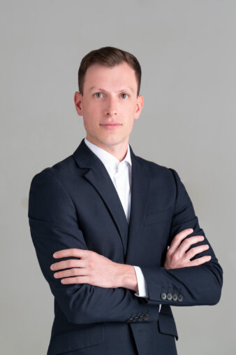 Un uomo in abito scuro e camicia bianca si trova su uno sfondo semplice, con le braccia incrociate, e guarda direttamente la telecamera con un'espressione neutra.