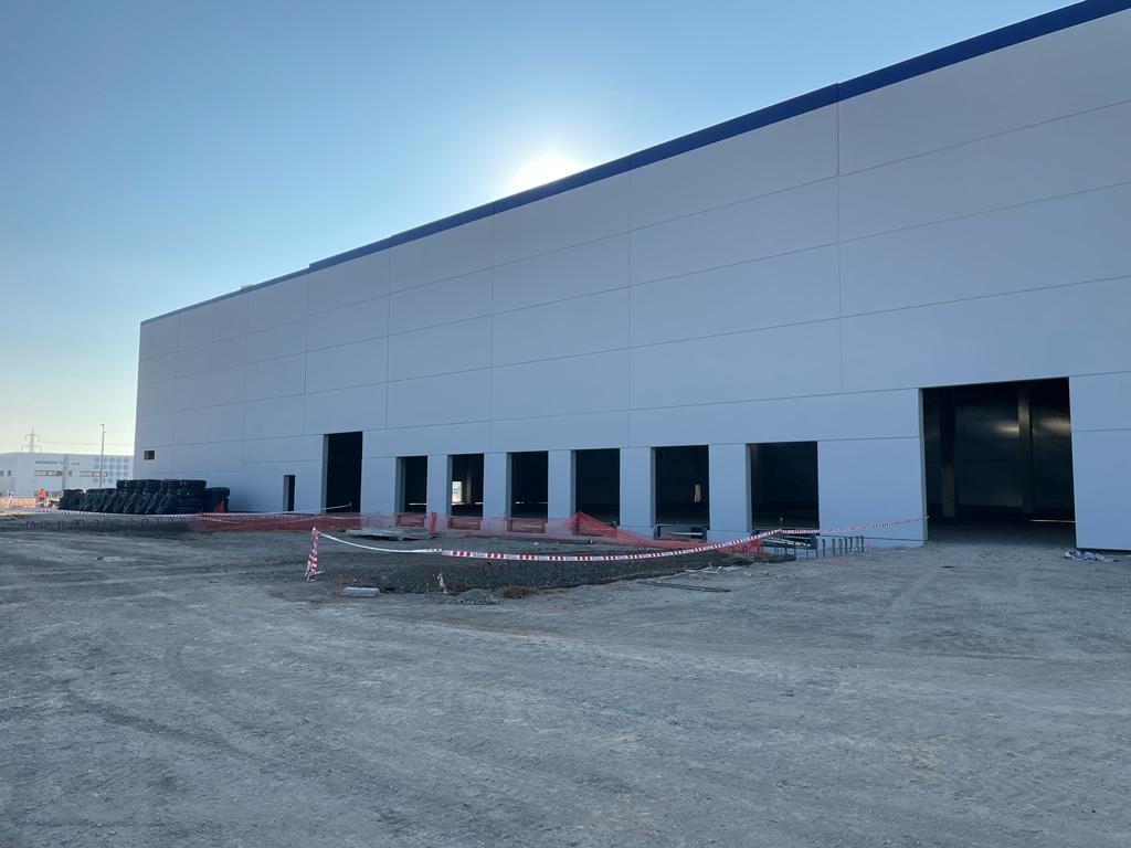 Grande edificio industriale bianco in costruzione, con portoni aperti, materiali da costruzione e un'area recintata sul davanti, su un terreno sterrato.