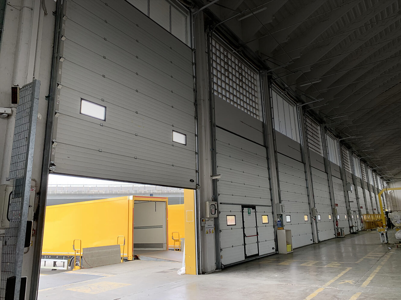 Grande banchina di carico industriale con diverse porte basculanti chiuse e una porta aperta che mostra un camion giallo per le consegne appoggiato alla banchina per lo scarico o il carico.