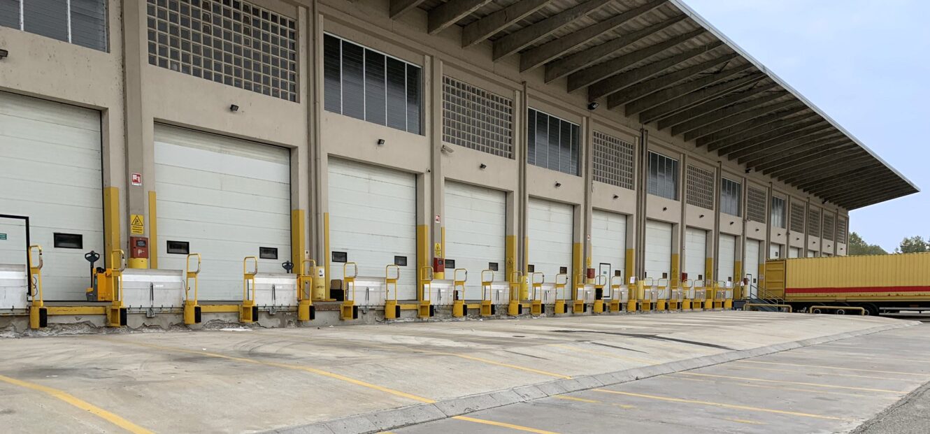 Una fila di porte di carico chiuse in un magazzino, con barriere di sicurezza gialle e un rimorchio vuoto parcheggiato su una banchina.