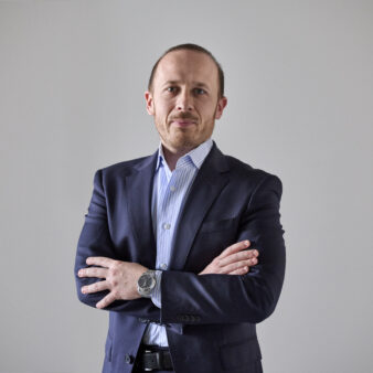 Un uomo in abito scuro e camicia a righe si trova su uno sfondo semplice con le braccia incrociate e guarda la telecamera.