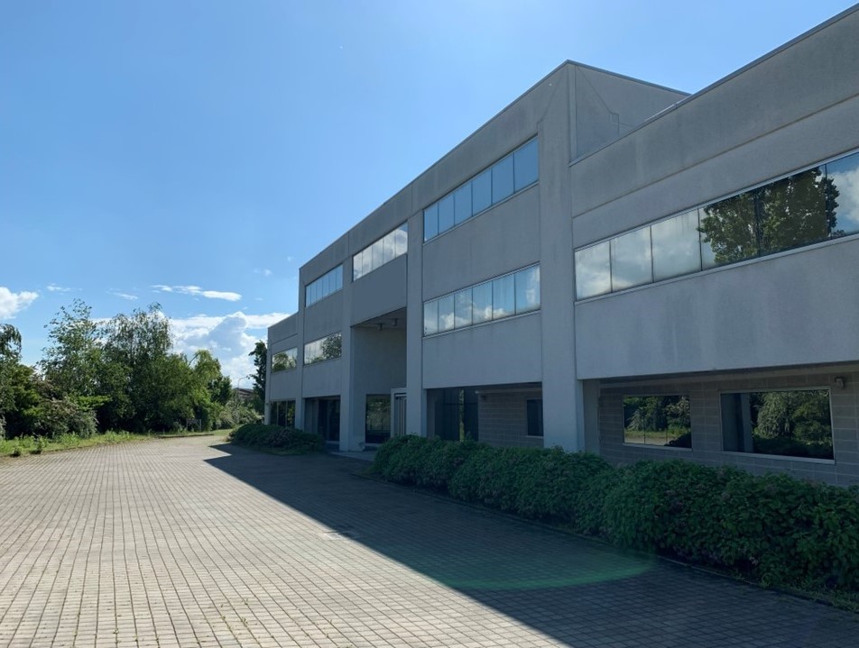 Un moderno edificio commerciale a due piani con ampie vetrate e un vialetto pavimentato, delimitato da cespugli verdi e circondato da alberi sotto un cielo azzurro e limpido.