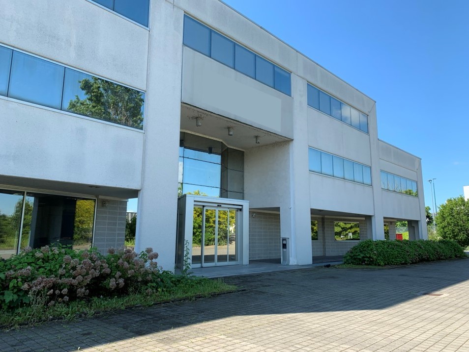 Edificio commerciale a due piani con ampie vetrate, porta d'ingresso in vetro e un vialetto pavimentato circondato da cespugli sotto un cielo sereno.