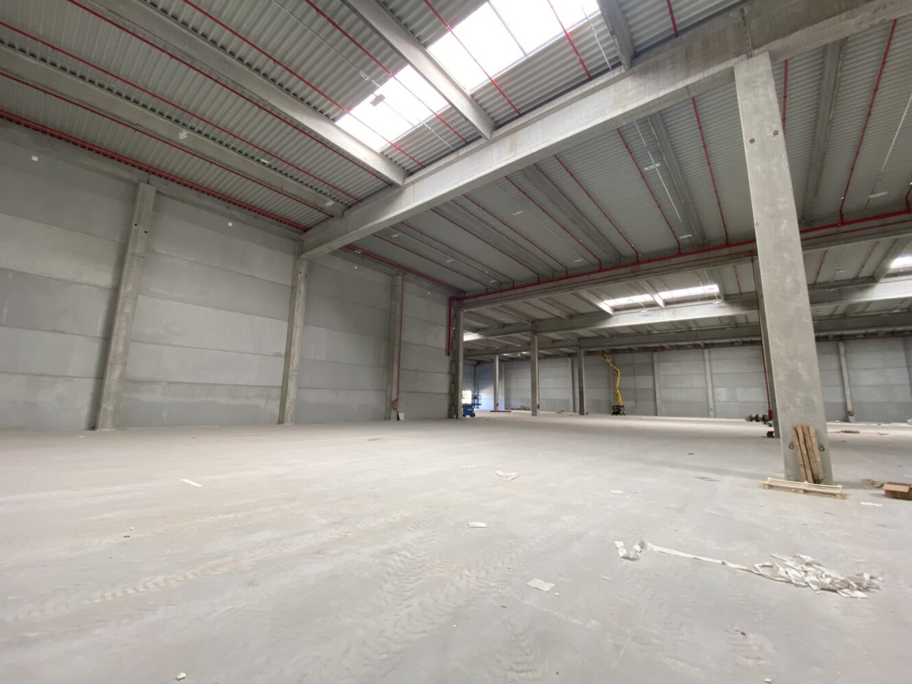 Grande magazzino industriale vuoto con soffitti alti, pavimenti in cemento e lucernari; visibili alcuni detriti sparsi e materiali da costruzione.