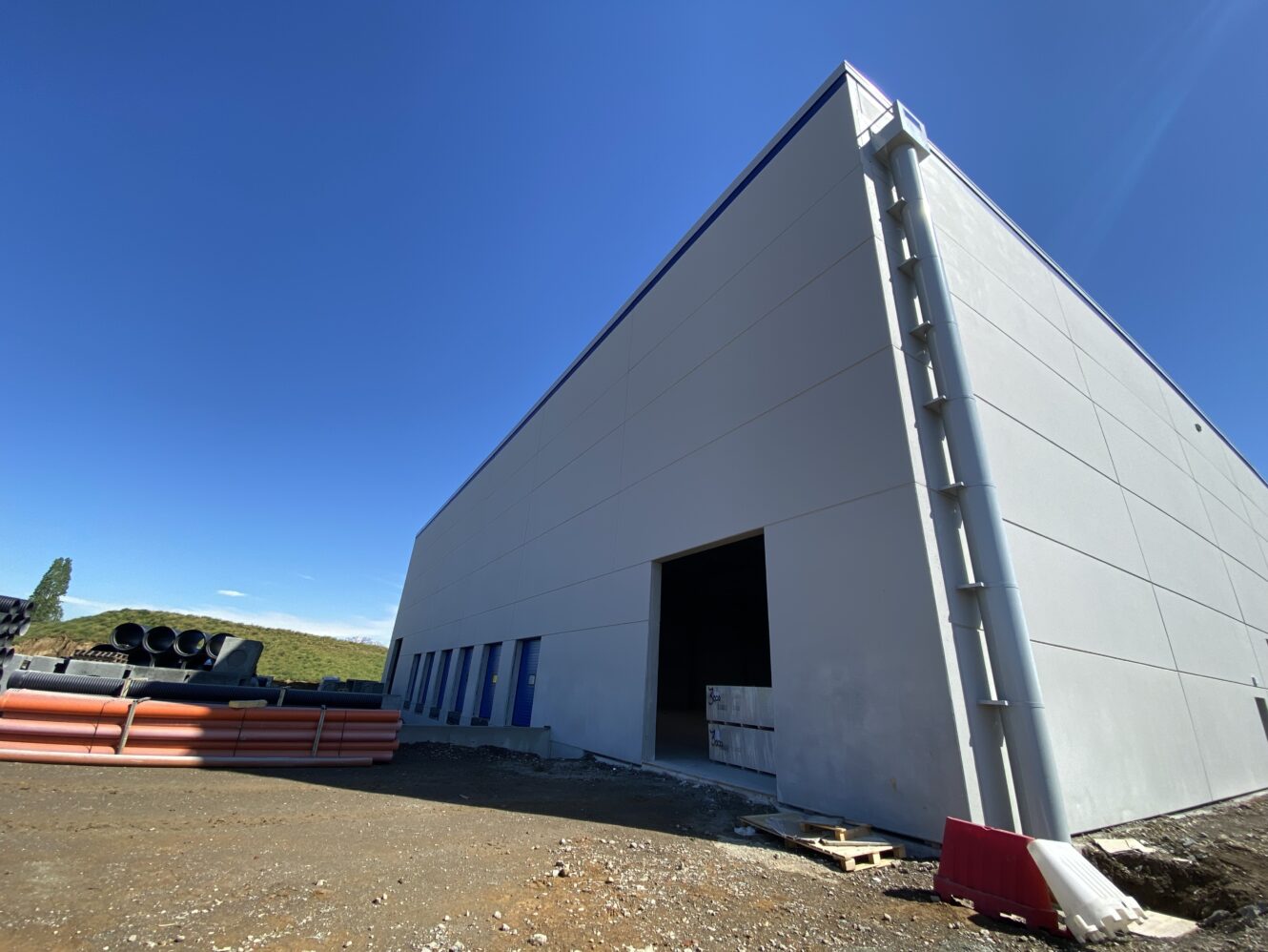 Un grande edificio industriale parzialmente costruito con un ingresso aperto, materiali da costruzione e tubi accatastati all'esterno sotto un cielo azzurro e limpido.