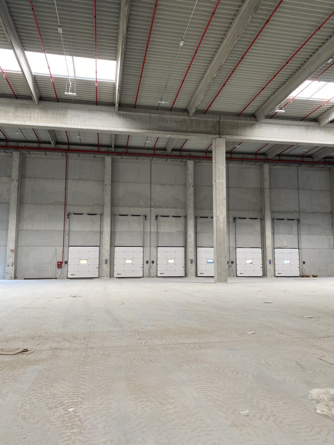 Interno di un magazzino industriale vuoto con pavimenti in cemento, soffitti alti e diverse grandi porte di carico lungo una parete.