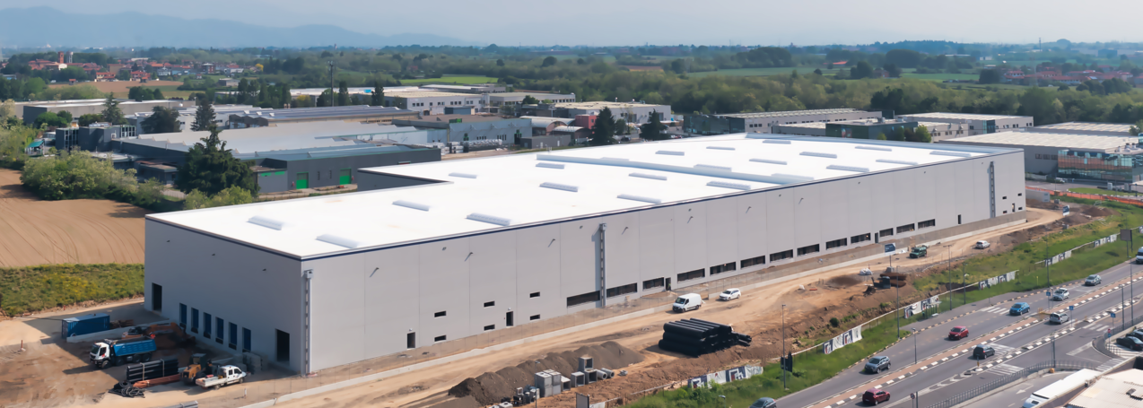 Un grande magazzino industriale rettangolare in costruzione, circondato da terra, materiali da costruzione e strade vicine con diversi veicoli.