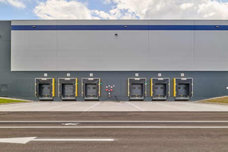 Sei banchine di carico di un moderno magazzino di distribuzione, ciascuna con porte basculanti e paraurti di protezione, viste da un parcheggio vuoto.