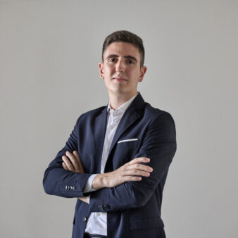 Un uomo in abito scuro con camicia bianca si trova su uno sfondo chiaro, con le braccia incrociate e rivolto in avanti con un'espressione neutra.