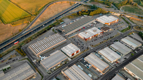 Vista aerea di un parco industriale con diversi grandi capannoni, parcheggi e strade intersecanti vicino a un'autostrada e a campi verdi.