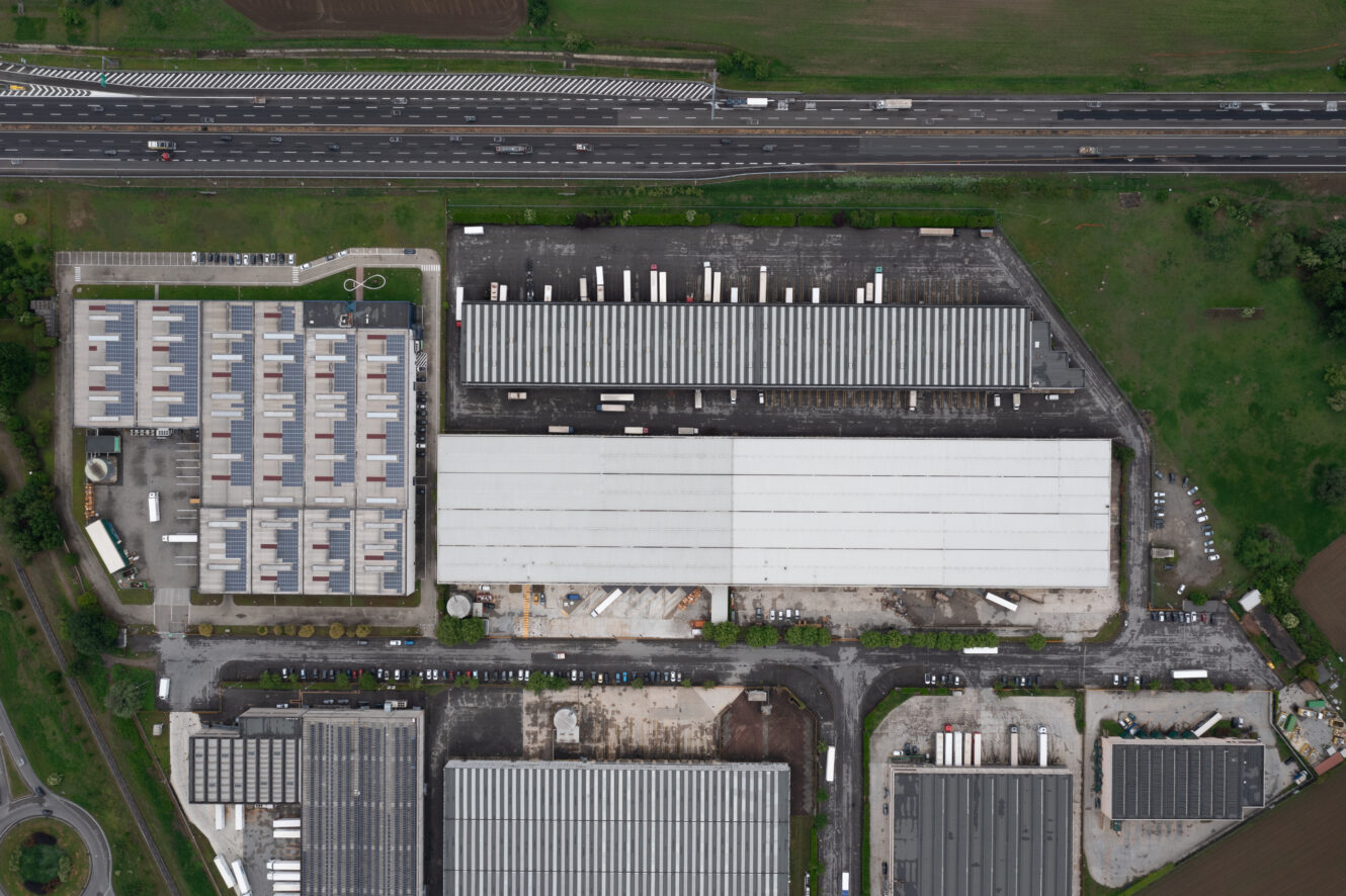 Veduta aerea di un grande impianto industriale con diversi magazzini, camion parcheggiati, container di stoccaggio e un'autostrada che corre lungo il bordo superiore dell'immagine.