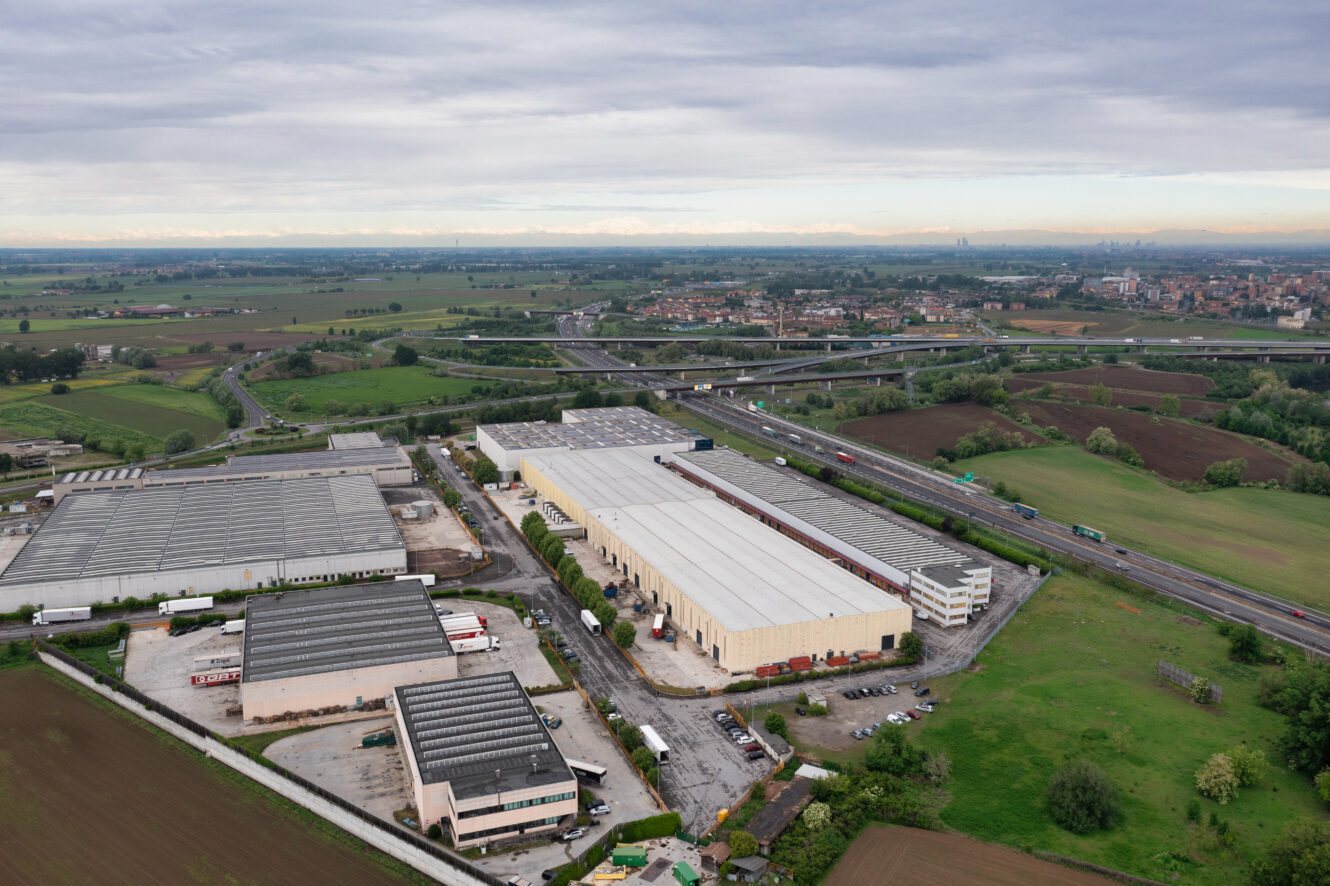 Veduta aerea di capannoni ed edifici industriali circondati da campi, con un'autostrada e un cavalcavia visibili sullo sfondo sotto un cielo nuvoloso.