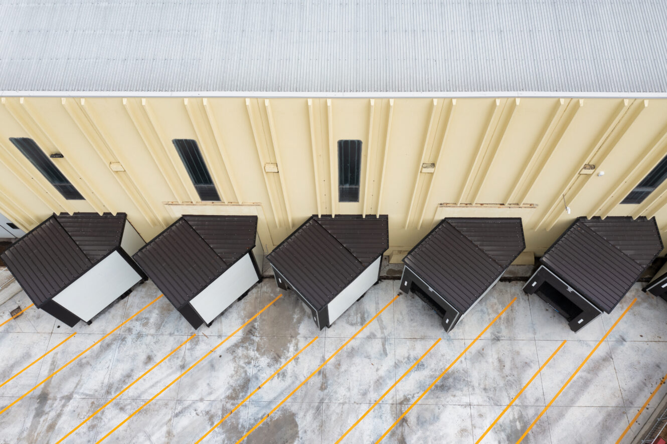 Veduta aerea di alcune piccole unità modulari bianche dal tetto scuro allineate accanto a un grande edificio industriale beige con parcheggi a strisce gialle.