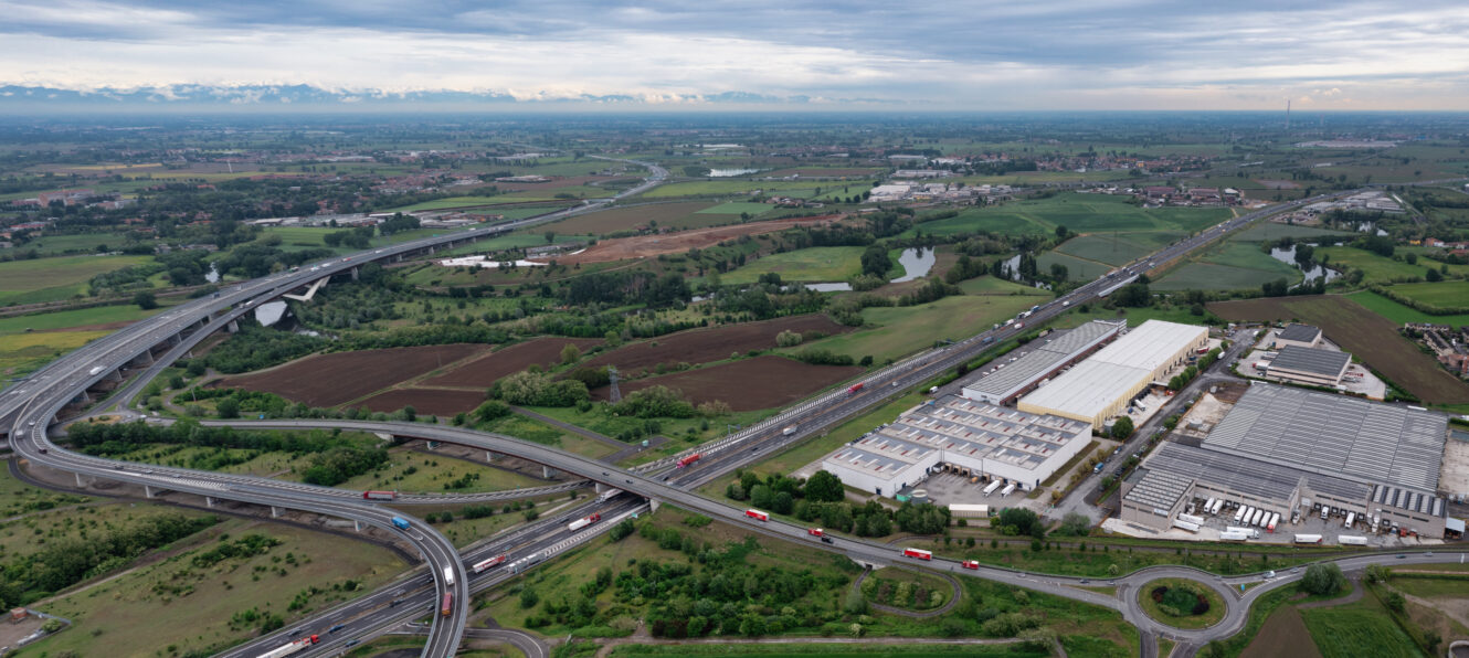 Vista aerea di uno svincolo autostradale vicino a edifici industriali e terreni agricoli, con più veicoli e campi verdi sotto un cielo nuvoloso.