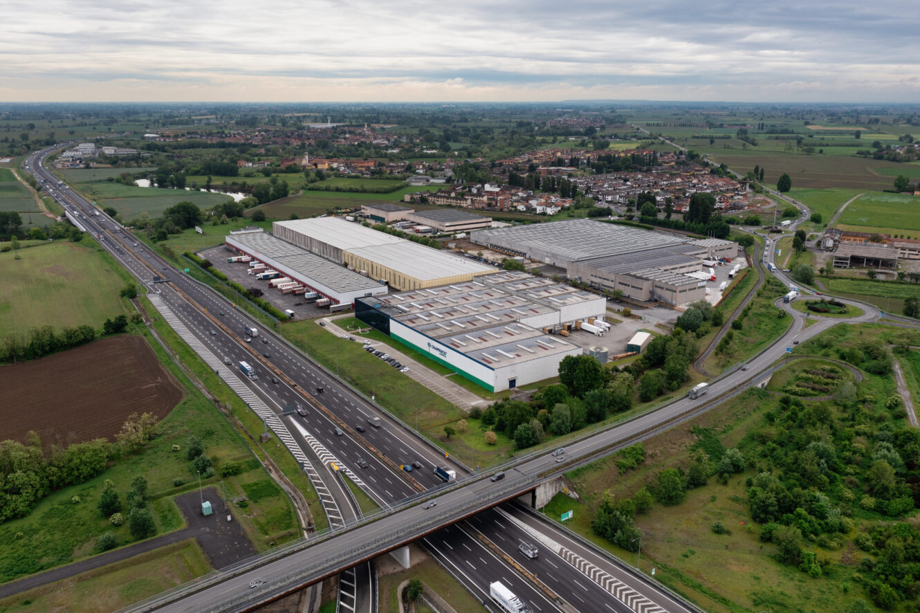 Veduta aerea di un complesso industriale vicino a uno svincolo autostradale, con camion parcheggiati e campi che circondano l'area sotto un cielo nuvoloso.