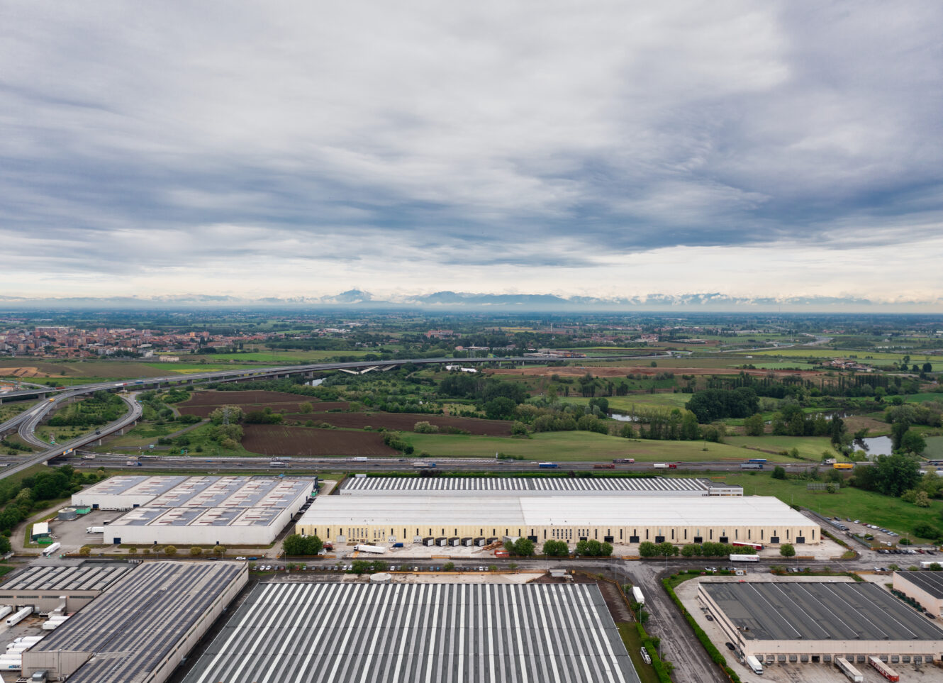 Veduta aerea di grandi edifici industriali con terreni agricoli, strade e città lontane sotto un cielo nuvoloso.