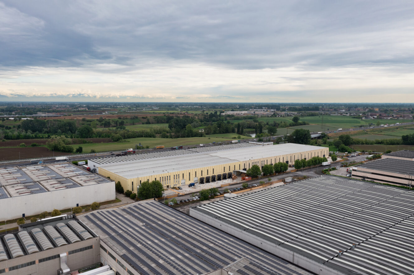 Veduta aerea di un'area industriale con grandi magazzini, circondata da campi verdi e da un cielo nuvoloso.