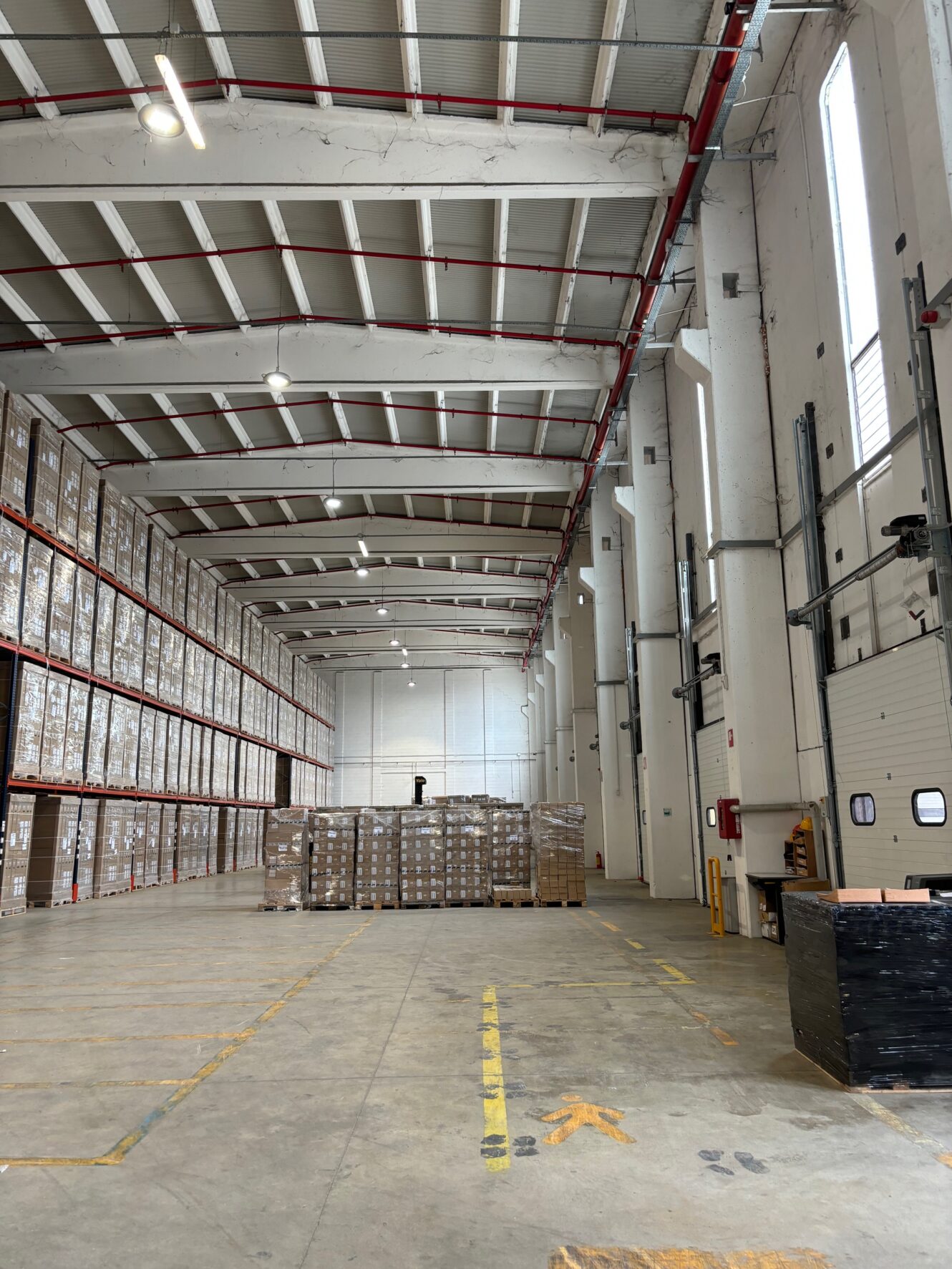 Interno di un grande magazzino con alti scaffali impilati di scatole sulla sinistra, un'area centrale con pallet di merci e un soffitto alto con luci sopraelevate.