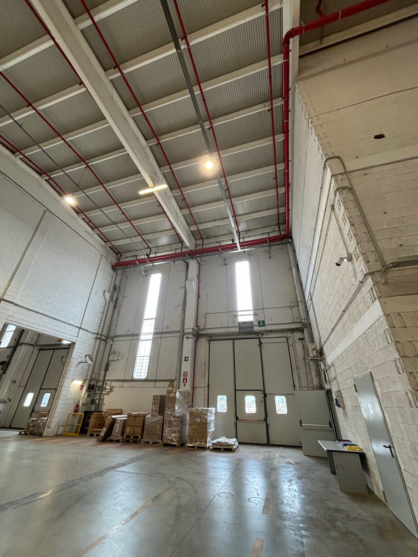 Ampio magazzino industriale con soffitti alti, pallet impilati e una scrivania vicino a grandi finestre e porte.