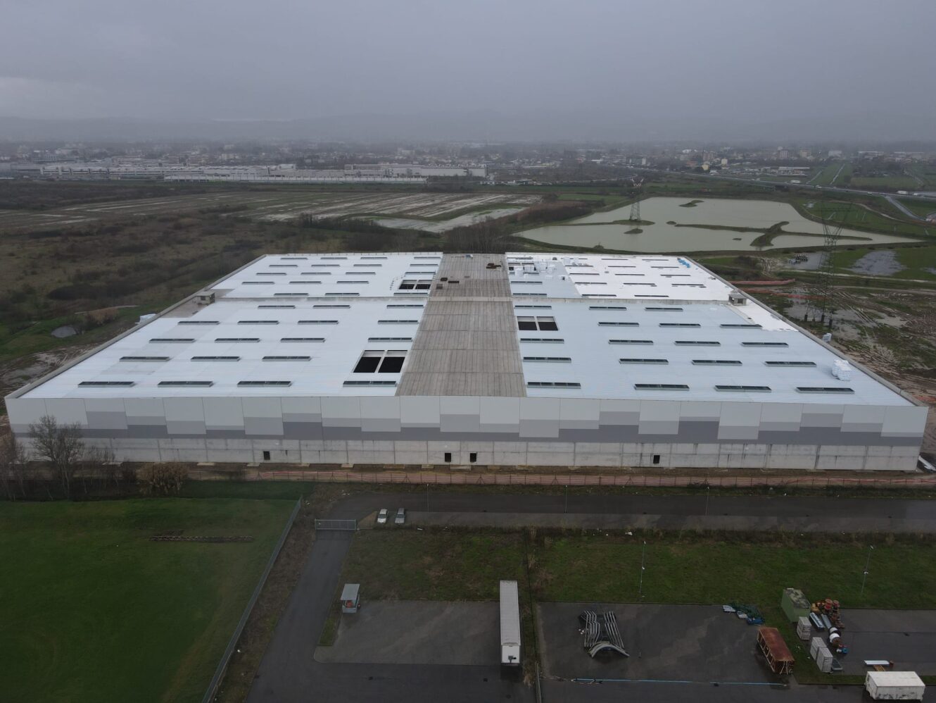 Un grande capannone industriale rettangolare con il tetto bianco si trova in un'area rurale circondata da campi sotto un cielo coperto.