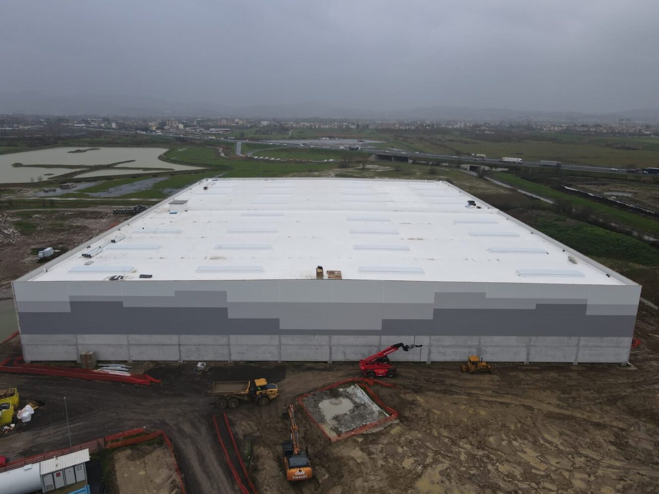 Un grande magazzino rettangolare in costruzione, con veicoli e attrezzature da cantiere su un terreno fangoso, campi e una città alle spalle.