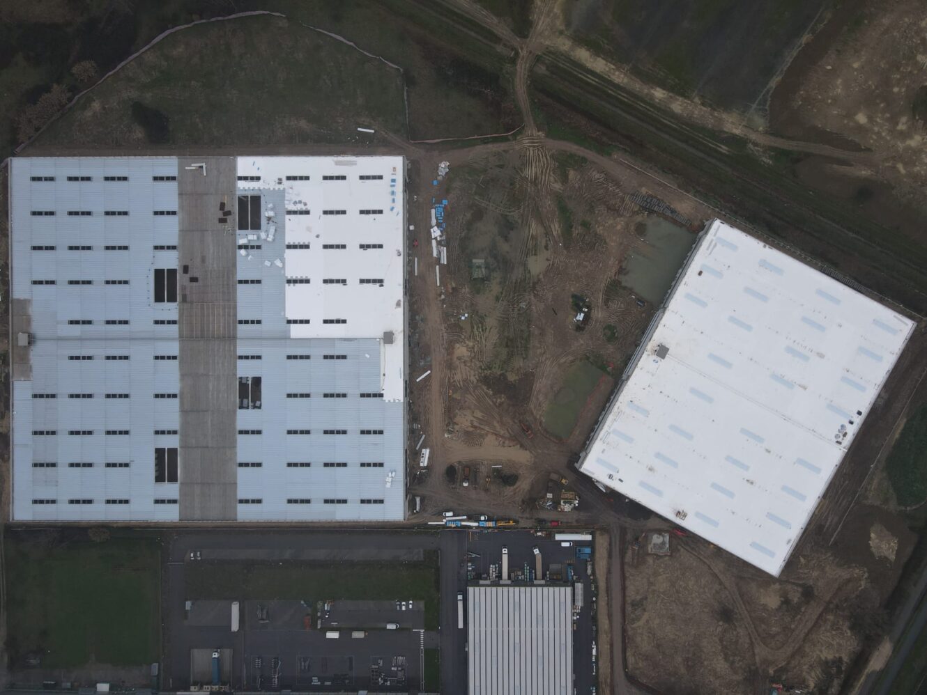 Veduta aerea di due grandi edifici industriali rettangolari con tetti bianchi, circondati da attrezzature edili, strade ed erba.