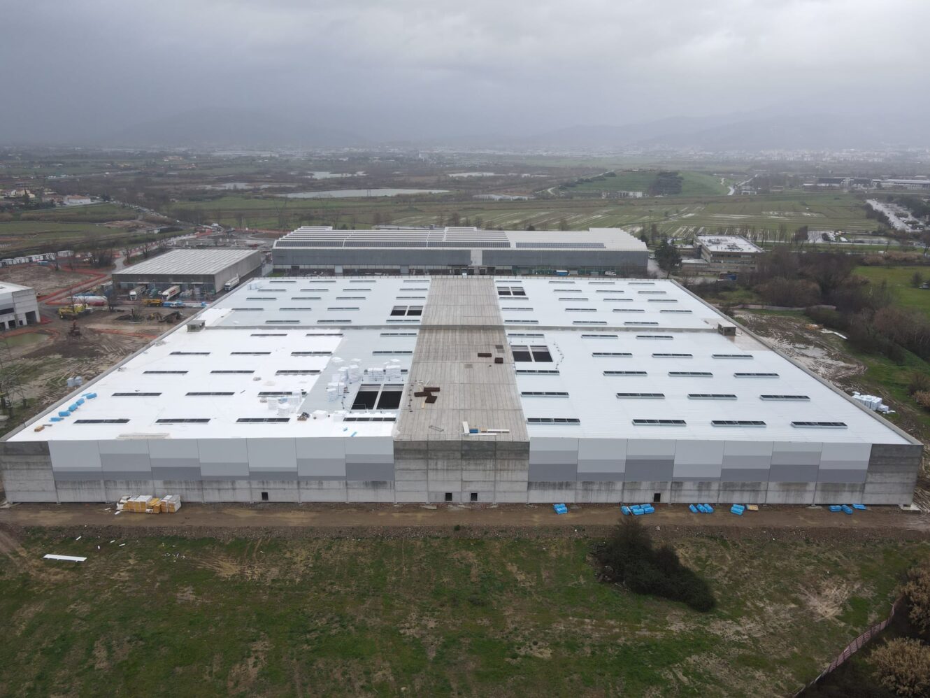 Veduta aerea di un grande capannone industriale in costruzione con tetto bianco, circondato da campi verdi e altri edifici.