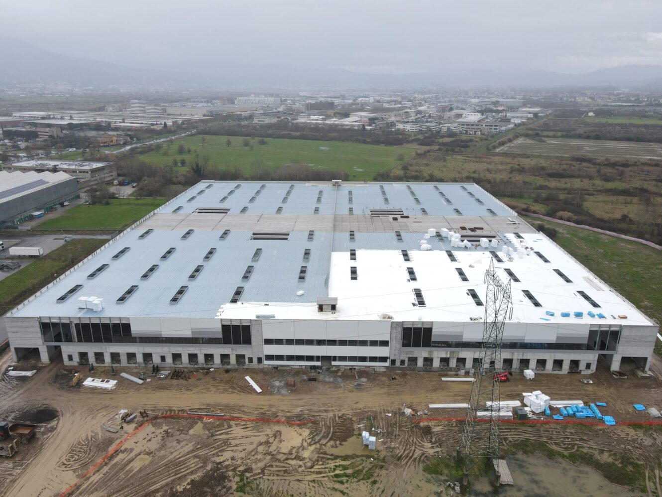 Veduta aerea di un grande capannone industriale in costruzione, materiali e attrezzature sparsi sul terreno, campi intorno.