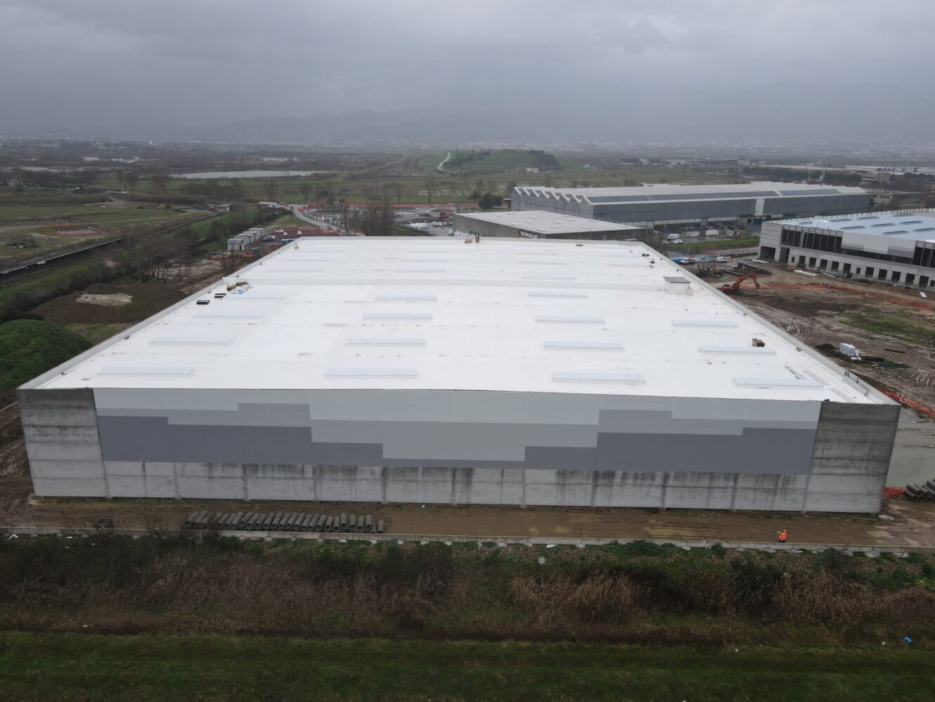 Grande magazzino industriale con tetto bianco e grigio, circondato da materiali da costruzione e macchinari in un'area semi-rurale sotto un cielo coperto.